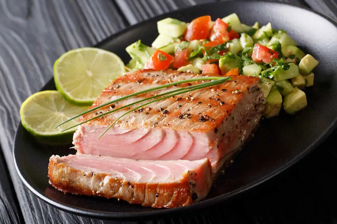 Grilled tuna steak served with fresh avocado and tomato salad, garnished with lime slices and chives.