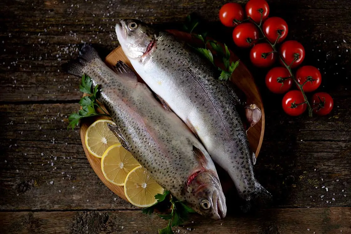 Two fresh whole trout fish placed on a wooden board with lemon slices, cherry tomatoes on the vine, and parsley.