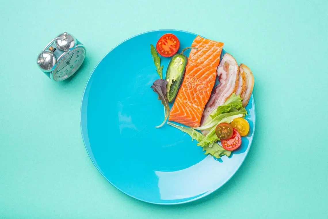 Blue plate with salmon, bacon, leafy greens, cherry tomatoes, and jalapeno slices next to an alarm clock, symbolizing intermittent fasting with omega-3-rich with fish high in omega 3s.