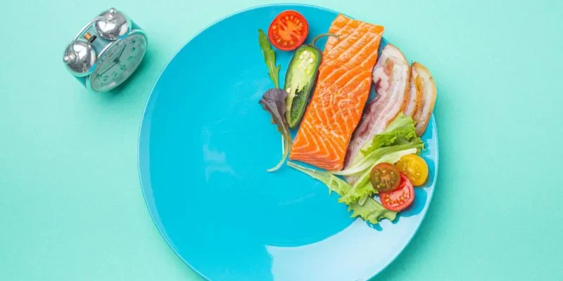 Blue plate with salmon, bacon, leafy greens, cherry tomatoes, and jalapeno slices next to an alarm clock, symbolizing intermittent fasting with omega-3-rich with fish high in omega 3s.