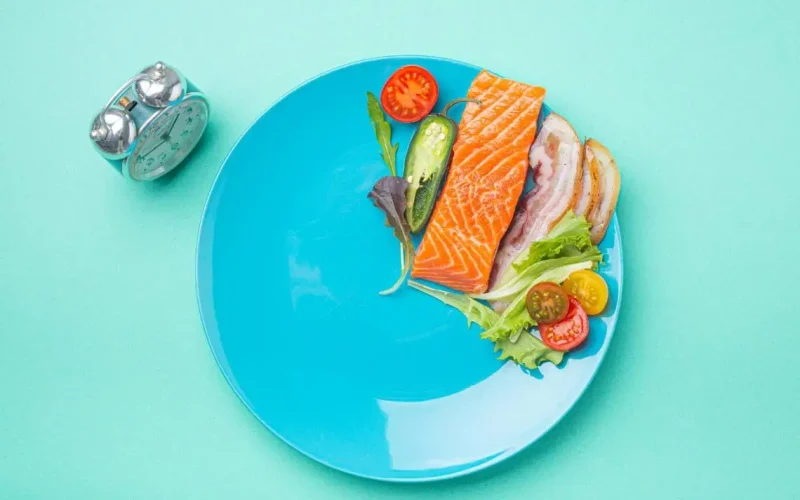 Blue plate with salmon, bacon, leafy greens, cherry tomatoes, and jalapeno slices next to an alarm clock, symbolizing intermittent fasting with omega-3-rich with fish high in omega 3s.